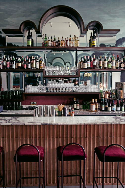 Bancone bar in marmo con rivestimento ondulato, scaffali in legno con specchi e bottiglie di liquore, sedie imbottite in velluto bordeaux.